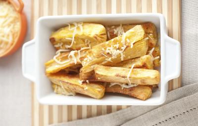 Fotografia em tons de bege em uma mesa de madeira com uma toalha bege e uma travessa retangular branca com as mandiocas assadas com parmesão dentro. Fotografia em tons de bege em uma mesa de madeira com uma toalha bege e uma travessa retangular branca com as mandiocas assadas com parmesão dentro.