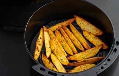 Fotografia de batatas assadas e temperadas, cortadas em meia lua, dentro da cestinha da Air Fryer preta sobre uma mesa escura.