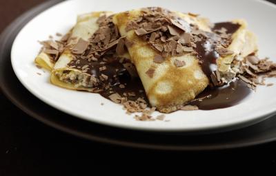 Fotografia de algumas panquecas, uma por cima da outra, recheadas e cobertas de brigadeiro, e por cima, raspas de chocolate. As panquecas estão em um prato branco que está sobre um sousplat marrom escuro. Fotografia de algumas panquecas, uma por cima da outra, recheadas e cobertas de brigadeiro, e por cima, raspas de chocolate. As panquecas estão em um prato branco que está sobre um sousplat marrom escuro.