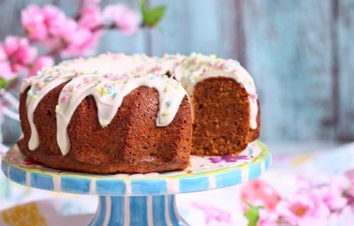 Fotografia de um bolo bundt elegantemente decorado, coberto com glacê branco e confeitos em tons pastel de estrelas e bolinhas. O bolo, de cor dourada e fofinho, está em um suporte listrado azul e branco, com um fundo de flores de cerejeira.