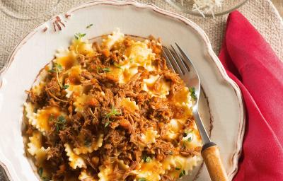Fotografia em tons de bege em uma mesa de madeira com um guardanapo de pano vermelho ao lado, um prato redondo fundo branco com detalhes vermelhos e o farfalle com ragu de carne dentro dele.