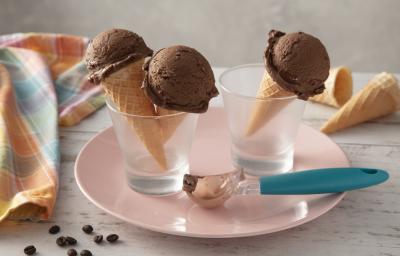 Fotografia em tons de azul e rosa de uma bancada, ao centro um prato rosa com dois copos, dentro deles casquinhas de sorvetes com uma bola do sorvete de café com chocolate. Sobre o prato, um boleador de sorvete com cabo azul.