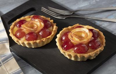 Fotografia em tons claros com uma toalha cinza listrada. Ao centro há um prato retangular preto com 2 tortas de frutas e ao fundo um par de garfos. Fotografia em tons claros com uma toalha cinza listrada. Ao centro há um prato retangular preto com 2 tortas de frutas e ao fundo um par de garfos.