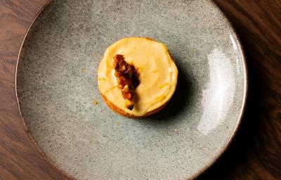 Fotografia em tons de marrom com um prato cinza ao centro. Dentro do prato existe um pequeno bolo de massa branca recheado com um creme de cor amarelada e coberto com castanhas caramelizadas.