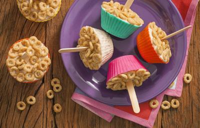 fotografia tirada de uma bancada marrom que contém um prato redondo roxo com 4 forminhas nas cores rosa, vermelho, branco e verde e todas contém um geladinho coberto com cereal e um palito de sorvete ao centro de cada um. fotografia tirada de uma bancada marrom que contém um prato redondo roxo com 4 forminhas nas cores rosa, vermelho, branco e verde e todas contém um geladinho coberto com cereal e um palito de sorvete ao centro de cada um.