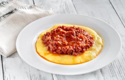 fotografia em tons de branco, amarelo e vermelho de uma bancada branca vista de cima. Contém um prato redondo branco com polenta, e por cima um molho bolonhesa. fotografia em tons de branco, amarelo e vermelho de uma bancada branca vista de cima. Contém um prato redondo branco com polenta, e por cima um molho bolonhesa.