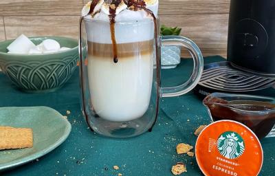Fotografia em tons de verde em uma bancada com um pano verde, uma xícara de vidro grande com a bebida de leite e café preparada dentro dela. Decorada com marshmallow, calda de chocolate e biscoito triturado. Fotografia em tons de verde em uma bancada com um pano verde, uma xícara de vidro grande com a bebida de leite e café preparada dentro dela. Decorada com marshmallow, calda de chocolate e biscoito triturado.