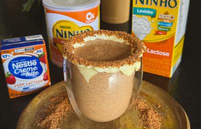 Fotografia de uma xícara de vidro com uma bebida de ninho com o creme de ninho na borda e ninho tostado também na borda e no prato raso em que a xícara está apoiada. O prato está sobre uma mesa preta, ao lado do prato tem produtos zero lactose nestlé Fotografia de uma xícara de vidro com uma bebida de ninho com o creme de ninho na borda e ninho tostado também na borda e no prato raso em que a xícara está apoiada. O prato está sobre uma mesa preta, ao lado do prato tem produtos zero lactose nestlé