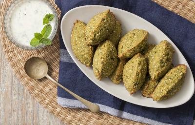 Fotografia em tons de verde, azul e branco, ao centro uma travessa branca com os bolinhos de falafel, ao lado para acompanhar um potinho de vidro com o molho de iogurte e uma folhinhas verdes para decorar. Abaixo está um guardanapo azul. Fotografia em tons de verde, azul e branco, ao centro uma travessa branca com os bolinhos de falafel, ao lado para acompanhar um potinho de vidro com o molho de iogurte e uma folhinhas verdes para decorar. Abaixo está um guardanapo azul.