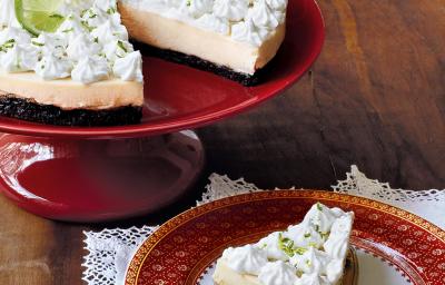 Fotografia em tons de vermelho e branco, com boleira vermelha contendo uma torta com creme e cobertura brancos, ao lado prato branco com borda vermelha com um pedaço de torta sob uma espátula e sobre guardanapo branco, tudo sobre bancada de madeira. Fotografia em tons de vermelho e branco, com boleira vermelha contendo uma torta com creme e cobertura brancos, ao lado prato branco com borda vermelha com um pedaço de torta sob uma espátula e sobre guardanapo branco, tudo sobre bancada de madeira.