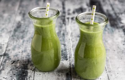 Fotografia de duas garrafas de vidro pequenas com um refresco de salsão, e em cada garrafinha há um canudo branco com bolinhas verdes. Os refrescos estão sobre uma mesa de madeira branca com detalhes pretos.