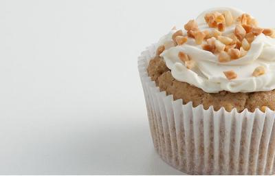 Fotografia em tons de bege em uma mesa branca com uma forminha de doce grande branca com o cupcake de massa integral dentro dele. Decorado com castanhas-de-caju.