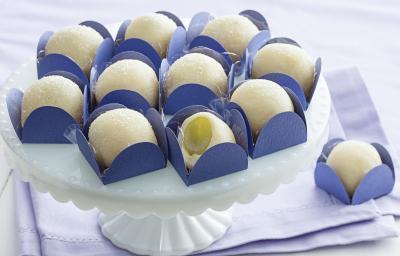 Fotografia em tons de branco e azul de uma bancada branca com paninho azul, sobre ele uma bailarina branca com docinhos de brigadeiro branco com uva verde dentro de forminhas quadradas azuis.
