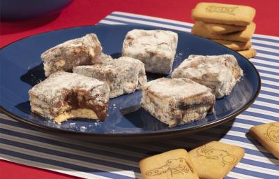 Fotografia em tons de azul e vermelho de uma bancada vermelha com paninho listrado de azul e branco, sobre ele um prato azul escuro com palha italiana de biscoito passatempo. Ao fundo biscoitos passatempo leite espalhados.