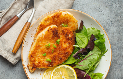 Imagem mostra um prato com duas fatias de frango empanado, folhas verdes e limões sicilianos amarelos. Garfo, faca ao lado.