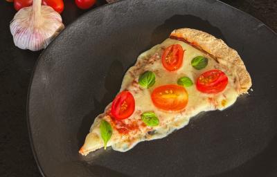 Fotografia de um prato raso preto sobre uma bancada preta. Dentro do prato tem uma fatia de pizza com queijo, tomate e manjericão. Ao lado do prato tem duas cabeças de alho inteiras com vários tomates cereja e um vidro de azeite. Fotografia de um prato raso preto sobre uma bancada preta. Dentro do prato tem uma fatia de pizza com queijo, tomate e manjericão. Ao lado do prato tem duas cabeças de alho inteiras com vários tomates cereja e um vidro de azeite.