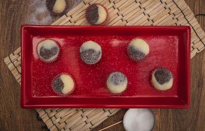 Fotografia em tons de vermelho em uma bancada de madeira escura, um jogo americano de palha, um recipiente retangular vermelho com brigadeiros mesclados de brigadeiro branco e preto em forminhas de vermelho. Fotografia em tons de vermelho em uma bancada de madeira escura, um jogo americano de palha, um recipiente retangular vermelho com brigadeiros mesclados de brigadeiro branco e preto em forminhas de vermelho.