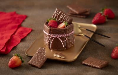 Foto de uma bancada escura com alguns tecidos vermelhos, morangos e biscoitos cobertos de chocolate espalhados. Ao centro há um prato retangular dourado com a receita pronta montada e decorada, acompanhada de duas colheres douradas e pretas Foto de uma bancada escura com alguns tecidos vermelhos, morangos e biscoitos cobertos de chocolate espalhados. Ao centro há um prato retangular dourado com a receita pronta montada e decorada, acompanhada de duas colheres douradas e pretas