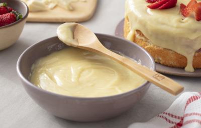 Fotografia em tons de vermelho em uma bancada de madeira cinza, um pano branco com listras vermelhas, um recipiente cinza com a ganache de chocolate branco. Ao fundo, um bolo coberto com a ganache e decorado com morangos.