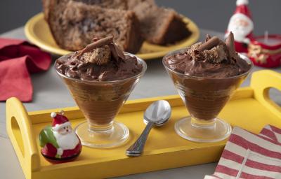 Fotografia em tons de amarelo e vermelho em uma bancada de madeira com decoração natalina, ao centro uma bandeja amarela com duas taças recheadas com chocolate e panettone e um paninho vermelho listrado ao lado.