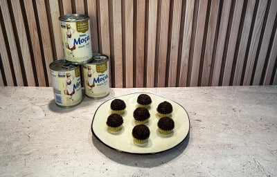 Fotografia em tons de bege em uma bancada, com latas de Leite Moça ao fundo e um prato ao centro com brigadeiros.