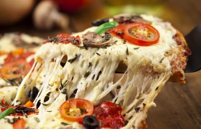 Fotografia de uma pizza com o queijo puxando e bem recheada, como queijo, azeitona, tomates e ricota. A pizza está sobre uma tábua de madeira de toma escuro, e ao fundo, cebola, tomate e champignon.