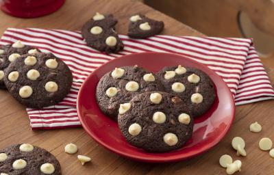 Fotografia em tons de vermelho em uma bancada de madeira com um pano branco com listras vermelhas, um prato vermelho ao centro com três cookies de chocolate com gotas de chocolate branco. Espalhados pela mesa, vários cookies de chocolate.