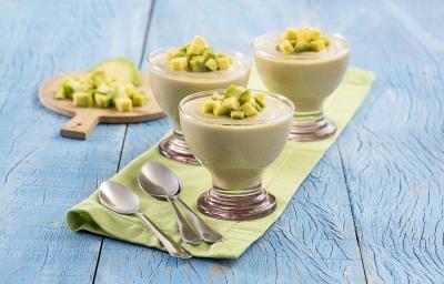 Foto da receita de mousse de abacate disposta em três taças de vidro com abacate em cubos para decorar, estão em cima de uma mesa azul com um pano verde separando, em cima do pano duas colheres prateadas. Ao fundo, abacate em cubos numa tábua de madeira Foto da receita de mousse de abacate disposta em três taças de vidro com abacate em cubos para decorar, estão em cima de uma mesa azul com um pano verde separando, em cima do pano duas colheres prateadas. Ao fundo, abacate em cubos numa tábua de madeira