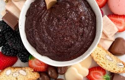 Fotografia em tons de vermelho e rosa em uma bancada com uma toalha colorida. Ao centro, um prato redondo fundo com o creme de chocolate de brownie com frutas, castanhas, frutas secas e pães ao redor dele. Fotografia em tons de vermelho e rosa em uma bancada com uma toalha colorida. Ao centro, um prato redondo fundo com o creme de chocolate de brownie com frutas, castanhas, frutas secas e pães ao redor dele.
