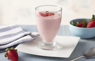 Fotografia em tons de azul e rosa de uma bancada com um prato quadrado, sobre ele um copo com o creme de morango e pedacinhos de morango. Ao redor um paninho listrado azul e branco e um potinho com morangos.
