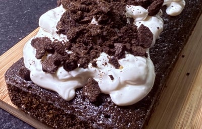 Fotografia de um bolo de negresco, assado na forma de bolo inglês, sobre uma tábua de madeira. O bolo está decorado com iogurte natural e biscoitos negresco quebrados. Fotografia de um bolo de negresco, assado na forma de bolo inglês, sobre uma tábua de madeira. O bolo está decorado com iogurte natural e biscoitos negresco quebrados.