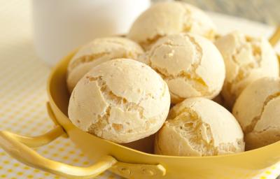 Fotografia em tons de amarelo em uma bancada de madeira esverdeada, um paninho branco com bolinhas amarelas, um recipiente amarelo com vários pães de queijo dentro dele. Ao fundo, uma xícara branca com café. Fotografia em tons de amarelo em uma bancada de madeira esverdeada, um paninho branco com bolinhas amarelas, um recipiente amarelo com vários pães de queijo dentro dele. Ao fundo, uma xícara branca com café.