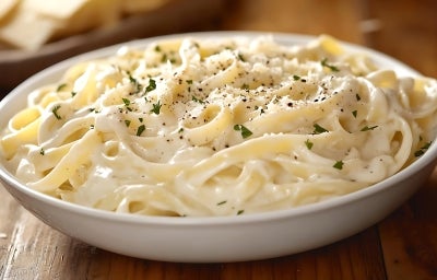 Fotografia em tons de branco com uma travessa branca ao centro. Em cima da travessa existe uma receita de macarrão com molho alfredo.