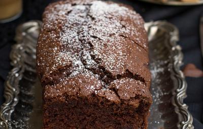 Fotografia em tons de marrom de uma bancada marrom vista de cima, contém uma bandeja de alumínio com um bolo inglês de chocolate com açúcar polvilhado por cima. Fotografia em tons de marrom de uma bancada marrom vista de cima, contém uma bandeja de alumínio com um bolo inglês de chocolate com açúcar polvilhado por cima.