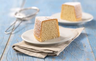 Foto da Receita de Mini Bolo de Baunilha. Observa-se duas fatias de bolo de baunilha, uma em cada prato branco sobre um guardanapo bege em uma superfície de madeira azul clara. Ao fundo, uma peneira com açúcar de confeiteiro de decoração.