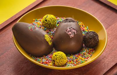 Fotografia em tons de amarelo, em uma bancada de madeira de cor marrom. Ao centro, um prato amarelo contendo os ovos de brigadeiro e granulados coloridos em volta. Ao fundo, alguns panos amarelos. Fotografia em tons de amarelo, em uma bancada de madeira de cor marrom. Ao centro, um prato amarelo contendo os ovos de brigadeiro e granulados coloridos em volta. Ao fundo, alguns panos amarelos.