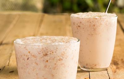 Fotografia em tons de marrom em uma mesa de madeira com dois copos de vidro com o frappé de amêndoas com coco dentro e um canudo de aço inox. Amêndoas espalhadas pela mesa.