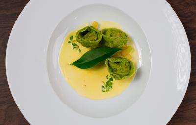 Fotografia em tons de branco com um prato branco ao centro. Em cima do prato existe uma receita de massa recheada de cor verde acompanhada de um molho de cor amarela. Fotografia em tons de branco com um prato branco ao centro. Em cima do prato existe uma receita de massa recheada de cor verde acompanhada de um molho de cor amarela.