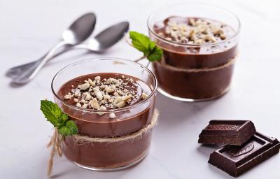 Fotografia de dois recipientes de vidro decorativos com pudim de café crocante com uma mini corda amarrando o pote e folhas de hortelã junto. Ao fundo, duas colheres de sobremesa, e ao lado, dois quadrados de chocolate amargo sobre uma mesa branca. Fotografia de dois recipientes de vidro decorativos com pudim de café crocante com uma mini corda amarrando o pote e folhas de hortelã junto. Ao fundo, duas colheres de sobremesa, e ao lado, dois quadrados de chocolate amargo sobre uma mesa branca.