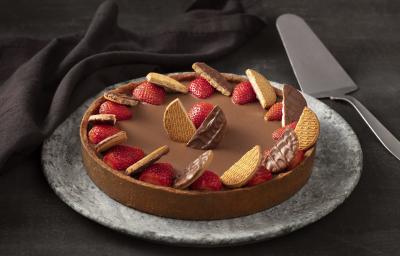 Fotografia em tons de preto, com fundo preto com paninho preto e prato cinza, com uma torta de chocolate, morangos e biscoitos Calipso e uma espátula prata. Fotografia em tons de preto, com fundo preto com paninho preto e prato cinza, com uma torta de chocolate, morangos e biscoitos Calipso e uma espátula prata.
