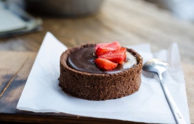 Fotografia em tons claros de uma mini torta de bombom de morango com base de chocolate sobre um papel dobrado e uma colher de sopa. Decorando a ganache, que está cobrindo a torta, tem alguns morangos decorando.