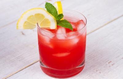 Fotografia em tons de vermelho em uma bancada de madeira de cor branca. Ao centro, um copo contendo a bebida pink lemonade.