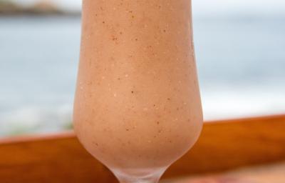 Fotografia de um copo de vidro, estilo taça, com uma bebida de suco de goiaba e neston sobre uma mesa de madeira com vista para a praia. Fotografia de um copo de vidro, estilo taça, com uma bebida de suco de goiaba e neston sobre uma mesa de madeira com vista para a praia.