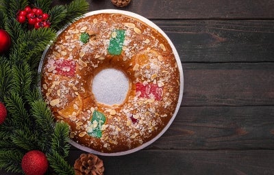 Fotografia de um bolo de reis enfeitado com frutas cristalizadas em vermelho, verde e laranja, polvilhado com açúcar e amêndoas. Colocado em um prato metálico, com ramos de pinheiro e enfeites vermelhos ao fundo.