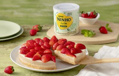 Fotografia em tons de verde e vermelho de uma bancada, sobre ela um prato redondo com uma sobremesa com base de biscoito, recheio de creme branco e morangos decorando. Ao fundo uma lata de NINHO Origem Vegetal e morangos.