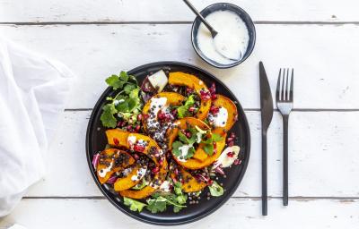 Fotografia vista de cima de uma salada de folhas com molho de laranja, pedaços de abóbora, sementes e um ramo de coentro em um prato raso preto. Ao lado tem talheres e um molho de iogurte em um pote pequeno de vidro azul, com uma colher apoiada. Fotografia vista de cima de uma salada de folhas com molho de laranja, pedaços de abóbora, sementes e um ramo de coentro em um prato raso preto. Ao lado tem talheres e um molho de iogurte em um pote pequeno de vidro azul, com uma colher apoiada.