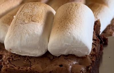 Fotografia de pedaços de brownie com marshmallow assados por cima sobre uma mesa branca.
