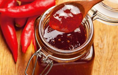 Fotografia de um recipiente de vidro decorativo com geleia de pimenta e laranja. O recipiente está sobre uma mesa de madeira, ao lado de várias pimentas dedo-de-moça. Fotografia de um recipiente de vidro decorativo com geleia de pimenta e laranja. O recipiente está sobre uma mesa de madeira, ao lado de várias pimentas dedo-de-moça.