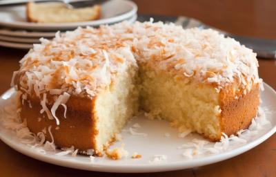 Fotografia de um bolo de coco com cobertura de coco e coco ralado apoiado em um prato branco raso. O bolo está com uma fatia grande cortada, e o prato está sobe uma mesa de madeira. Fotografia de um bolo de coco com cobertura de coco e coco ralado apoiado em um prato branco raso. O bolo está com uma fatia grande cortada, e o prato está sobe uma mesa de madeira.
