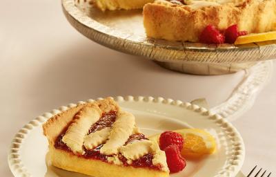 Fotografia em tons de bege em uma mesa de madeira, um guardanapo de pano bege e branco, um prato redondo branco com detalhes bege e uma fatia da crostata com recheio de laranja. Ao fundo, um prato redondo maior com a crostata inteira. Fotografia em tons de bege em uma mesa de madeira, um guardanapo de pano bege e branco, um prato redondo branco com detalhes bege e uma fatia da crostata com recheio de laranja. Ao fundo, um prato redondo maior com a crostata inteira.